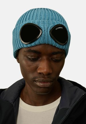 Berretto blu lavorato a maglia con dettagli in goggle neri, texture a costine e forma rotonda, indossato sopra una giacca con colletto e una camicia chiara.