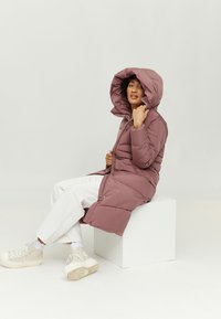 Langer, rosa, voluminöser Mantel mit großer Kapuze und Reißverschluss, mit taillierter Passform. Das Modell trägt weiße Hosen und helle Sneakers und sitzt auf einem Würfel.