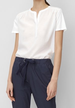 Vrouw draagt een witte blouse met korte mouwen en een gesplitste halslijn, gecombineerd met marineblauwe broek met trekkoord, tegen een effen achtergrond.