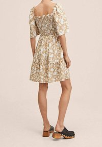 Robe à motif floral beige et blanc, avec un corsage froncé, des manches bouffantes et une jupe à volants. Associée à des sabots en bois noirs.