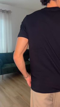 Osoba oblečená v černém tričku a béžových kalhotách stojí uvnitř vedle zelené pohovky, v pozadí je okno zakryté závěsem.