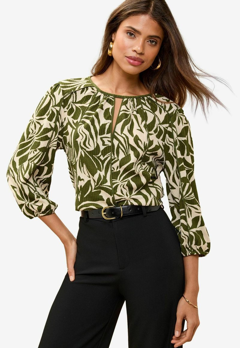 Groene en crèmekleurige blouse met patroon, lange poffende mouwen en een keyhole halslijn, gecombineerd met zwarte broek en een zwarte riem.