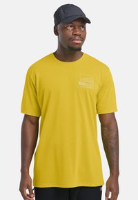 T-shirt giallo in cotone con colletto tondo e maniche corte, con un logo grafico bianco sul lato sinistro del petto. Cappello nero indossato sopra.