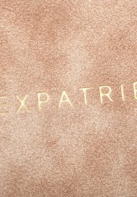 Rózsaszín, textúrált háttér, amelyen arany feliratok égetve olvasható "EXPATRIATE". A felület finoman szemcsés befejezéssel rendelkezik.