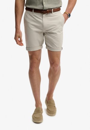 Kurze beige Shorts mit aufgerollten Bündchen, aus Baumwolle. Kombiniert mit einem braunen geflochtenen Gürtel und hellbraunen Slippern.