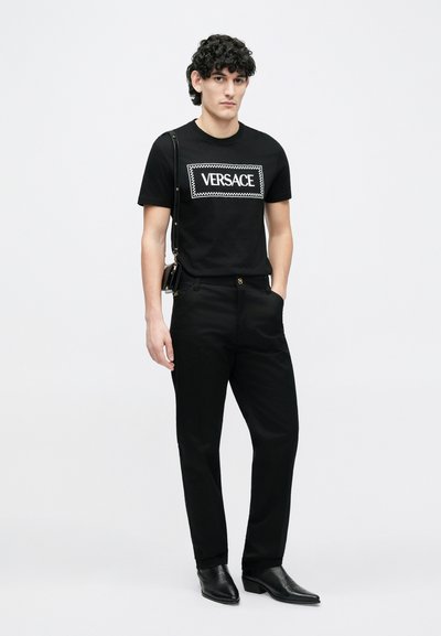 Fekete pamut póló fehér "VERSACE" logóval. Fekete nadrággal és fekete bőr csizmával párosítva, karcsúsított szabással és minimalista dizájnnal.