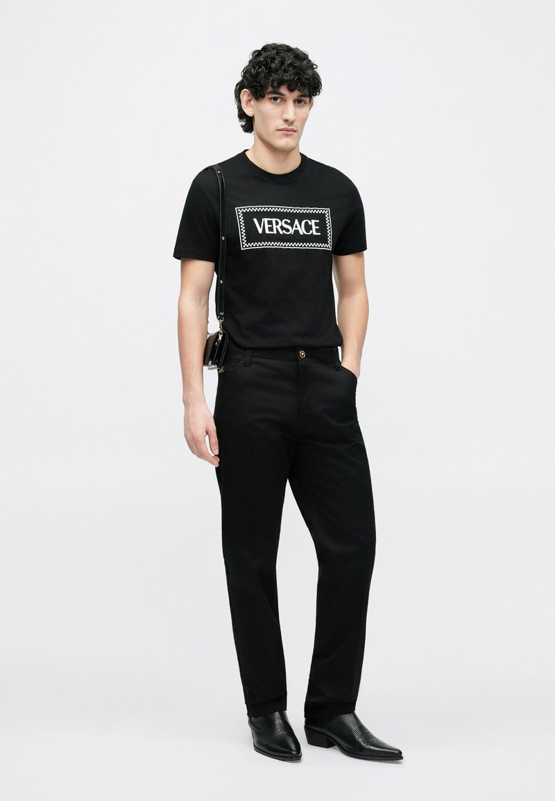 Svart bomullst-shirt med vitt "VERSACE"-logotyp. Kombinerad med svarta byxor och svarta läderstövlar, med en slimmad passform och minimalistisk design.