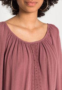 En mauve bluse med en rund halsudskæring, der har tekstureret stof og delikat blonde-detaljering i midten foran. Løs pasform.