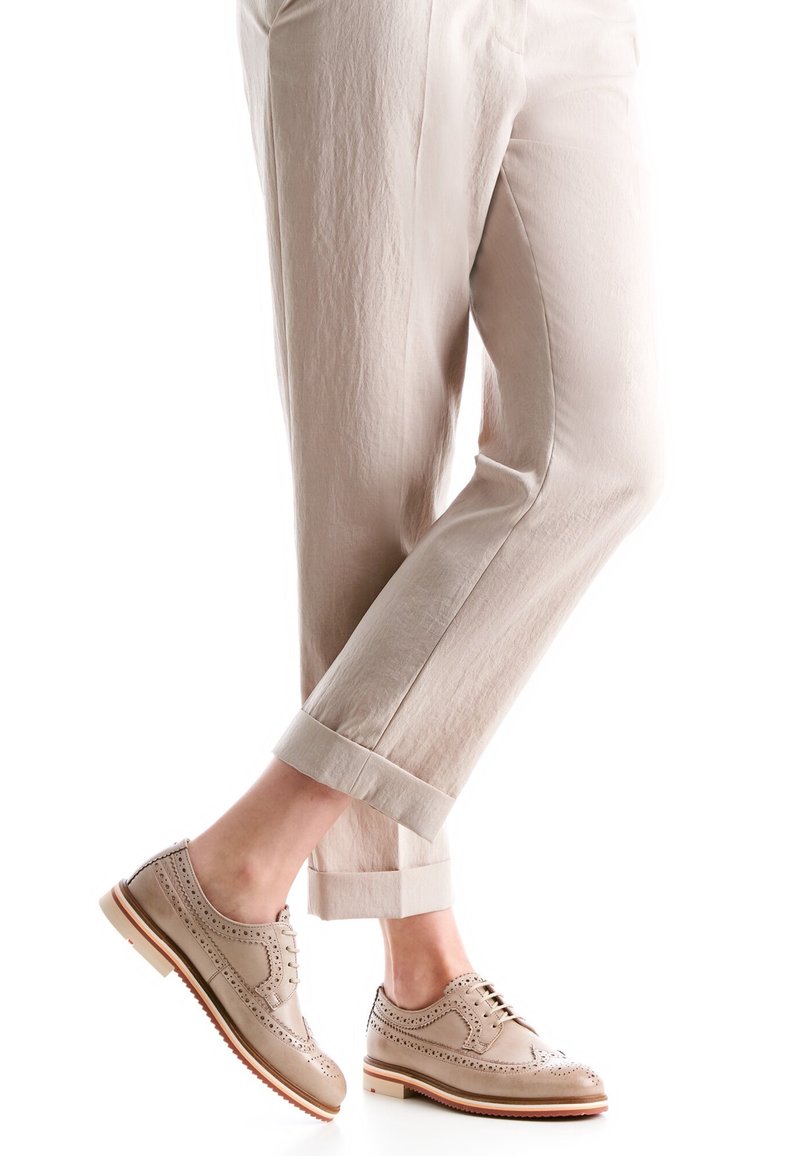 Pantalon beige retroussé associé à des chaussures brogues marron clair avec des détails perforés et des semelles en bois, portés par une personne debout sur fond blanc.