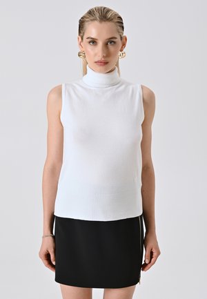 Blonde vrouw in een witte mouwloze coltrui, zwarte rok met zijrits en grote gouden ringoorbellen, staand tegen een effen achtergrond.