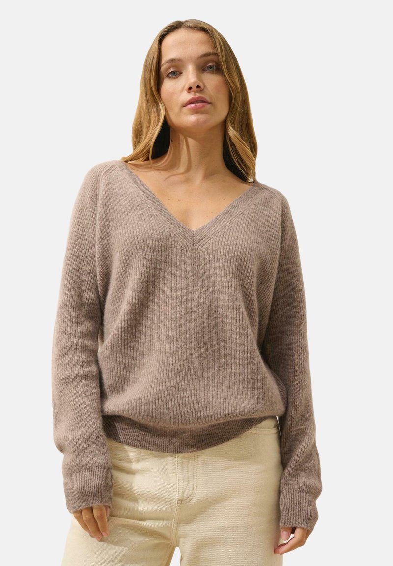 Jasnobrązowy sweter z prążkowanego materiału, z dekoltem w serek i długimi rękawami, charakteryzujący się luźnym krojem i miękką fakturą. Połączony z jasnymi spodniami.