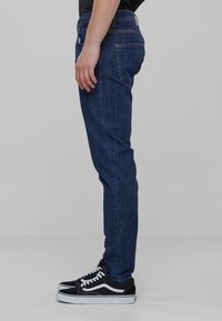 Kék farmernadrág, slim fit szabással, klasszikus öt zsebes dizájnnal és kontrasztos varrással, fekete-fehér sportcipővel párosítva.