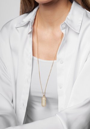 Collar de oro con un diseño de ondas texturizadas, colgado de una cadena delgada, llevado sobre una blusa blanca y debajo de una camisa de color claro.