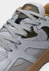 Λευκό και γκρι αθλητικό παπούτσι με ανοιχτό γκρι κορδόνια, επάνω μέρος από δίχτυ, καφέ σόλα και επικάλυψη με λογότυπο σε ελιά πράσινη απόχρωση στη γλώσσα.