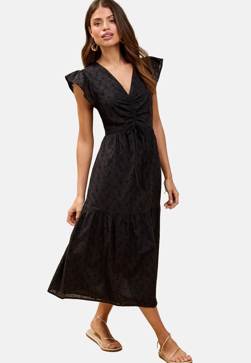 Robe noire midi avec un décolleté en V, un corsage froncé et des manches flottantes. Dotée d'une jupe en tiered et d'un motif floral subtil sur le tissu. Portée avec des sandales.