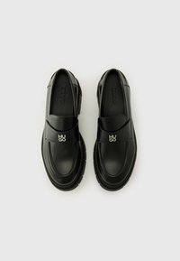 Black leather loafers with a rounded toe, prominent stitching, and a metal logo accent. Thick, textured sole for added grip.