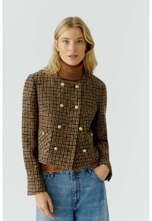 Blonde Frau, die eine braun karierte doppelt geknöpfte Jacke über einem braunen Rollkragenpullover trägt, mit blauen Jeans, posiert mit einer Hand in der Tasche.