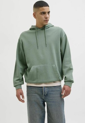 Sweat à capuche vert clair avec une poche avant, des cordons de serrage et des poignets côtelés, associé à un jean bleu. Texture douce, design décontracté.