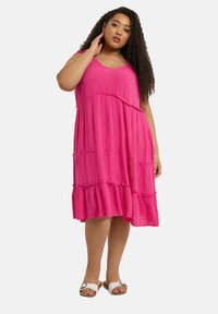 Rosa, lager-på-lager klänning med V-ringning och tunna axelremmar. Lättviktsmaterial med en texturerad yta, som faller över knäet. Bärs med vita sandaler.