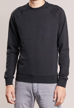 Homme portant un sweat-shirt noir Armani Jeans avec un logo embossé sur le devant et un pantalon gris clair sur un fond uni.
