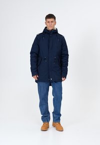 Temno modra obložena parka s kapuco, nastavljivimi vrvicami, dvema sprednjima žepoma in zadrgo, nošena čez modre kavbojke in rjave škornje.