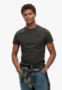 T-shirt grigio scuro a maniche corte con colletto rotondo e piccolo logo in rilievo, abbinato a jeans blu e una camicia a quadri legata in vita.