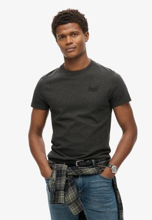T-shirt grigio scuro a maniche corte con colletto rotondo e piccolo logo in rilievo, abbinato a jeans blu e una camicia a quadri legata in vita.