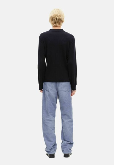 Personne aux cheveux blonds courts portant un haut noir à manches longues, un pantalon ample bleu clair et des chaussures noires, debout de dos sur un fond blanc.