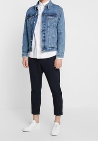 Denimová bunda ve středně modré barvě s kontrastním prošitím, s knoflíkovým zapínáním, dvěma náprsními kapsami; kombinováno s bílou košilí a tmavými zkrácenými kalhotami, bílé tenisky.