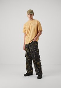 Lys oransje oversized t-skjorte, svarte kamuflasje cargo-bukser med lommer, olivengrønn caps, svarte sko, ensfarget bakgrunn.