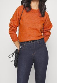 Pomarańczowy sweter z dzianiny z długimi rękawami w połączeniu z ciemnoniebieskimi dżinsami. Na jednym ramieniu zawieszona jest czarna pleciona torba.