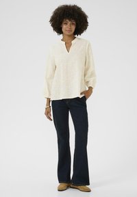 Šviesiai kreminė puloverio bluzonė su V formos iškirpte ir gėlėmis išsiuvinėtu ornamentu, derinama su tamsiai plačiomis džinsomis ir smėlio spalvos šlepetėmis.