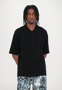 Polo shirt nero a maniche corte con colletto classico e pattina con tre bottoni, abbinato a pantaloni corti a fantasia con design floreale blu e bianco.