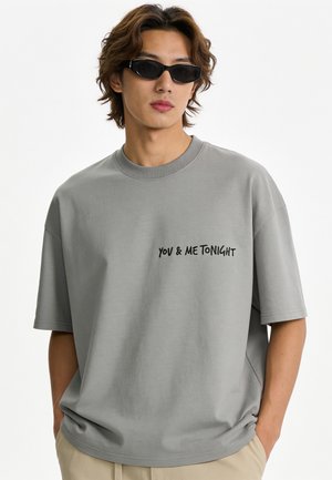 Junger Mann trägt schwarze Sonnenbrille und ein graues übergroßes T-Shirt mit dem Text "YOU & ME TONIGHT", Hände in den Taschen der beigen Hose.