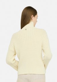 Maglione a coste cremoso con colletto alto, dotato di un dettaglio con logo argentato sul retro. La texture è morbida e leggermente elasticizzata.