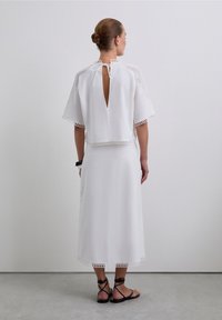 Robe blanche, jusqu'aux genoux, avec des manches courtes, présentant un dos à ouverture en forme de clé et un ourlet en dentelle à bords festonnés. Portée avec des sandales noires à lanières.