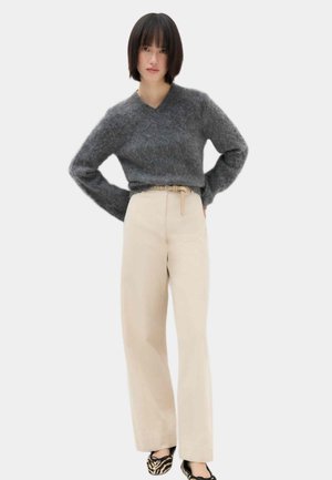 Junge Frau steht mit den Händen in der Hüfte, trägt einen grauen flauschigen Pullover, beige weite Hosen mit Gürtel und Leopardenmuster Ballerinas.