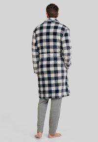 Bata de cuadros en blanco y negro hecha de una tela suave, con mangas largas, cinturón en la cintura y un diseño de longitud hasta la mitad de la pantorrilla. Llevada sobre pantalones grises.