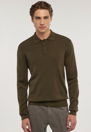 Joven con pelo corto y rizado, vistiendo una camiseta tipo polo de manga larga verde oliva y pantalones grises, de pie frente a un fondo liso.