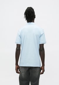 Svetlomodrá polokošeľa s krátkymi rukávmi a plochým golierom. Látka vyzerá hladká a ľahká, nosená s tmavosivými nohavicami.