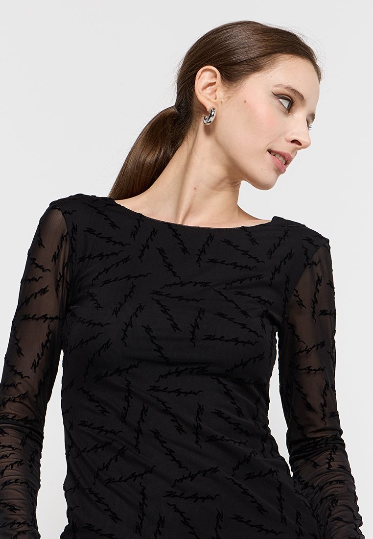 Mujer con cabello castaño en una coleta, usando aretes de aro de plata y una blusa negra transparente de manga larga con un patrón de texto diagonal.