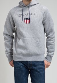 Felpa grigia con cappuccio a coulisse, tasca frontale e logo a scudo ricamato con strisce rosse e blu. Testo "GANT" sopra il logo.