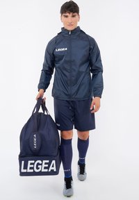 Giacca a vento blu con zip, polsini elasticizzati. Pantaloni corti blu e calze. Scarpe sportive nere e grigie. Grande borsa a tracolla blu con logo bianco.