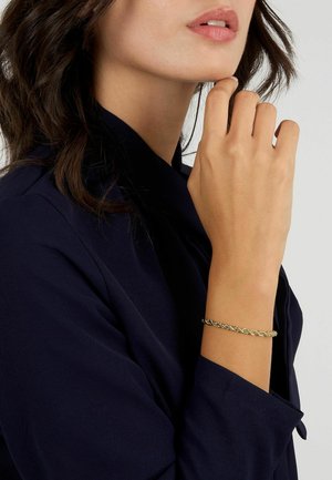 Bracciale oro intrecciato indossato al polso, con un design delicato e link intrecciati. Modella che indossa un top in tessuto blu navy.