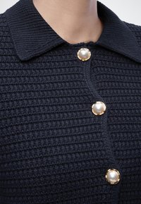 Σκούρο μπλε πλεκτή ζακέτα με υφή, με μεγάλα κουμπιά από μαργαριτάρια με χρυσά περίγραμμα και φαρδιά γιακά σε ανώτερο μέρος κορμού ενός ατόμου.