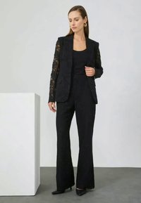 Black lace blazer with a tailored fit, paired with a black top and flared trousers. Smooth fabric with elegant lace accents on sleeves.