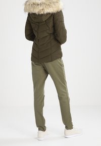 Chaqueta acolchada verde oliva con capucha forrada de piel y costuras en forma de chevron, combinada con pantalones a juego y zapatillas blancas, vista desde atrás.
