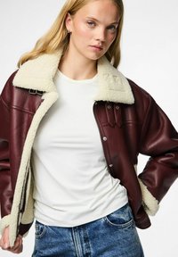 Giacca in pelle bordeaux con colletto in shearling crema, chiusura con zip e tasche frontali. Indossata sopra una camicia bianca con jeans blu.