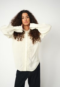 Cremfarbene Bluse aus leichtem Stoff, mit einer strukturierten Knopfleiste in der Mitte und voluminösen Ärmeln mit Bündchen.