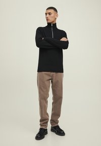 Pullover nero a costine con zip a un quarto, pantaloni beige a gamba dritta, scarpe nere con lacci, in posa in piedi su uno sfondo neutro.